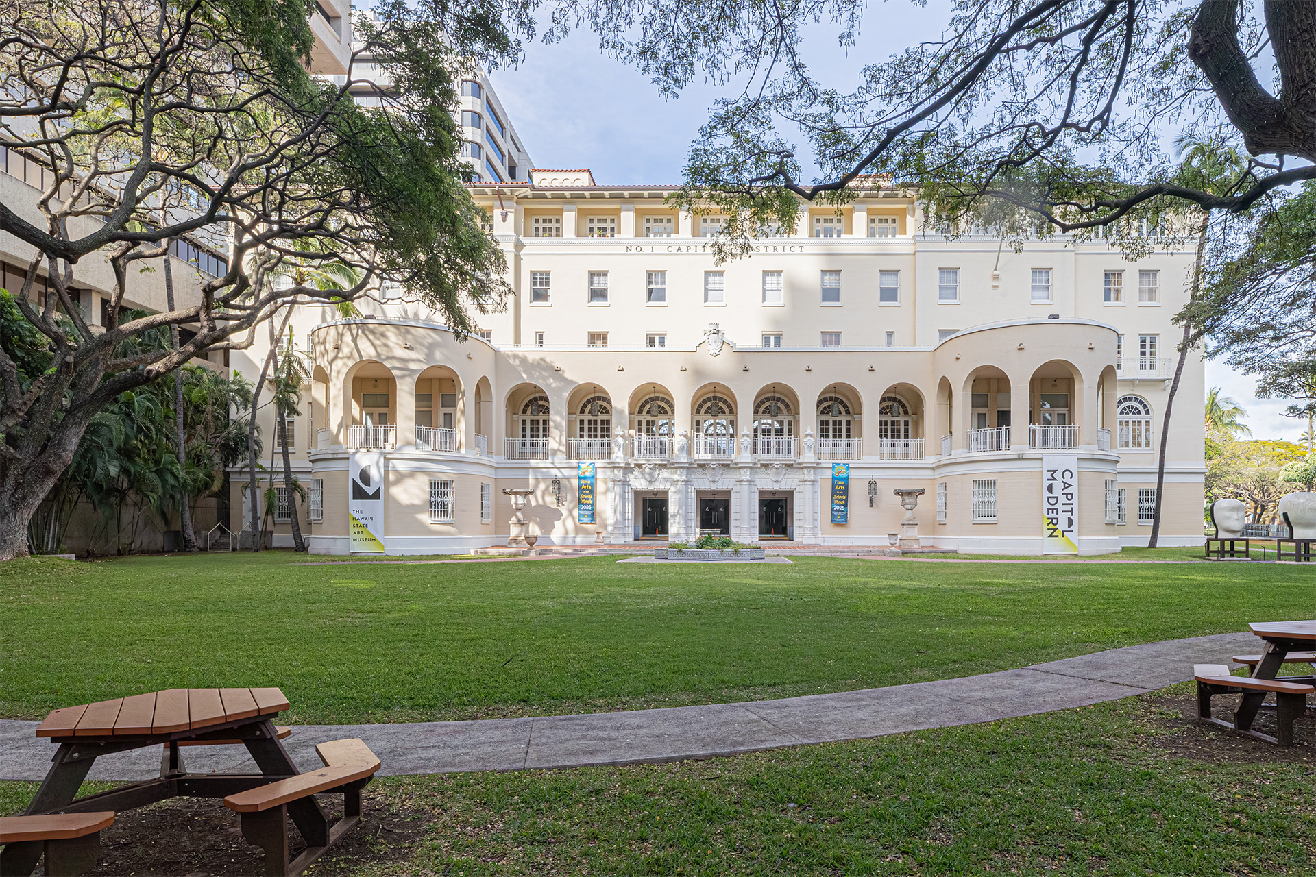 A large white building with arches and balconies surrounded by trees and a grassy area with picnic tables.