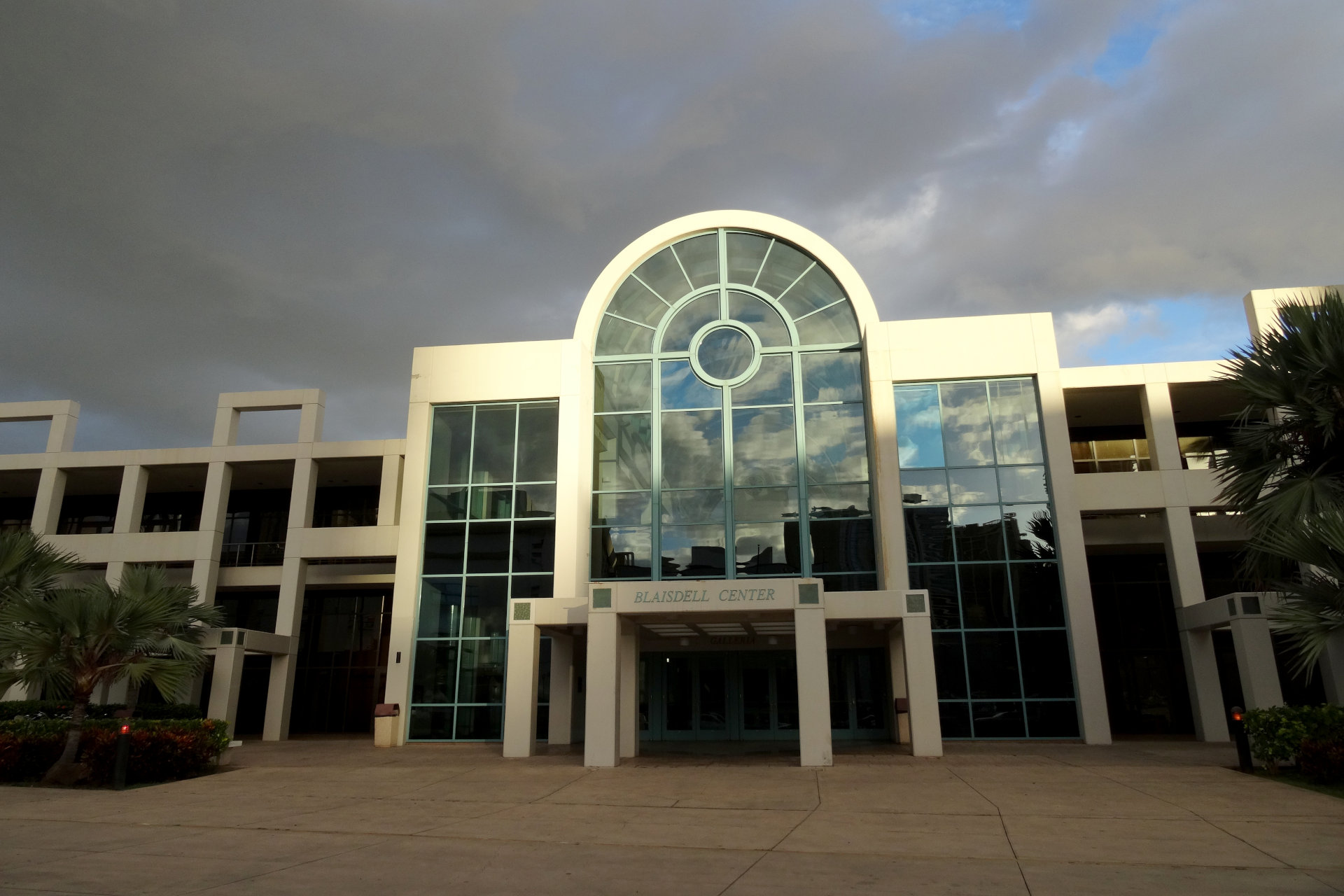 A large, modern building with a prominent arched glass section and palm trees in front.